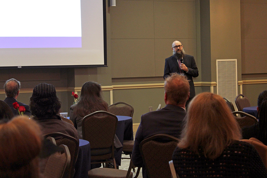 A male college professor presents data to over 100 people gathered in a large conference room.