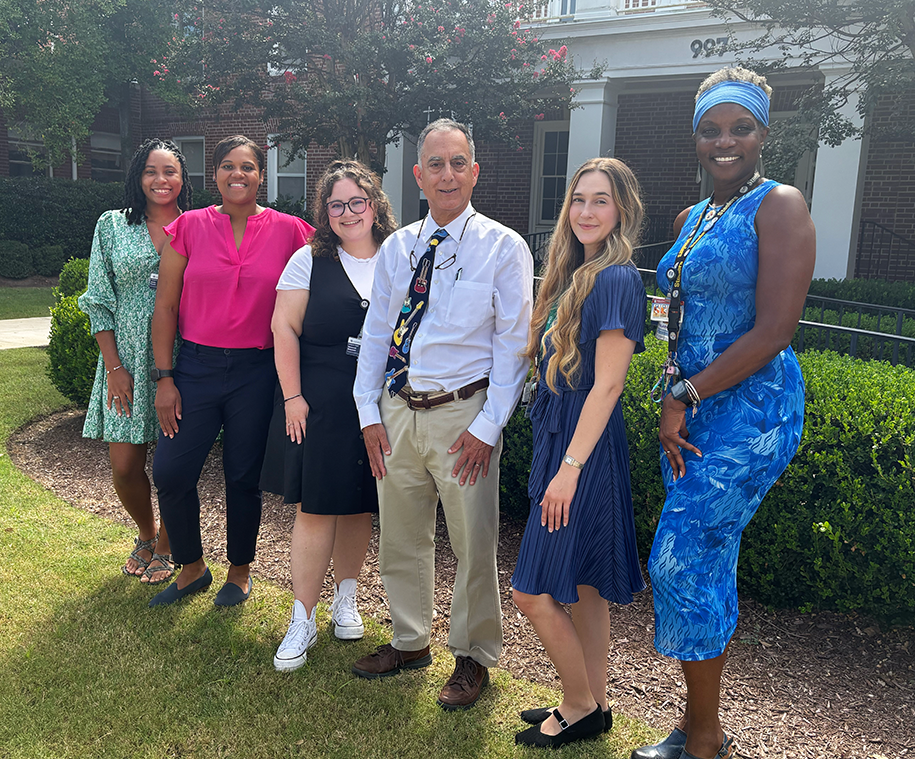 A group of six researchers at a medical college comprised of five women and one man stand outside in front of a building and smile at the camera.