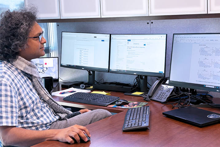 A man wearing a button down short sleeve shirt and a scarf sits in front of a computer and looks at the screen.