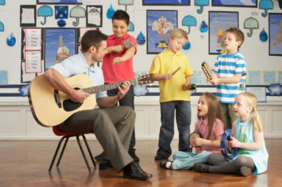 A male teacher accompanies elementary school singers by playing a guitar.