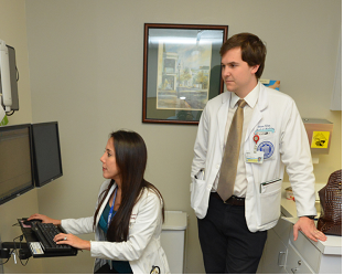 A female doctor sitting down and looking at a computer monitor, with a male resident doctor standing behind her and observingwhat she does.