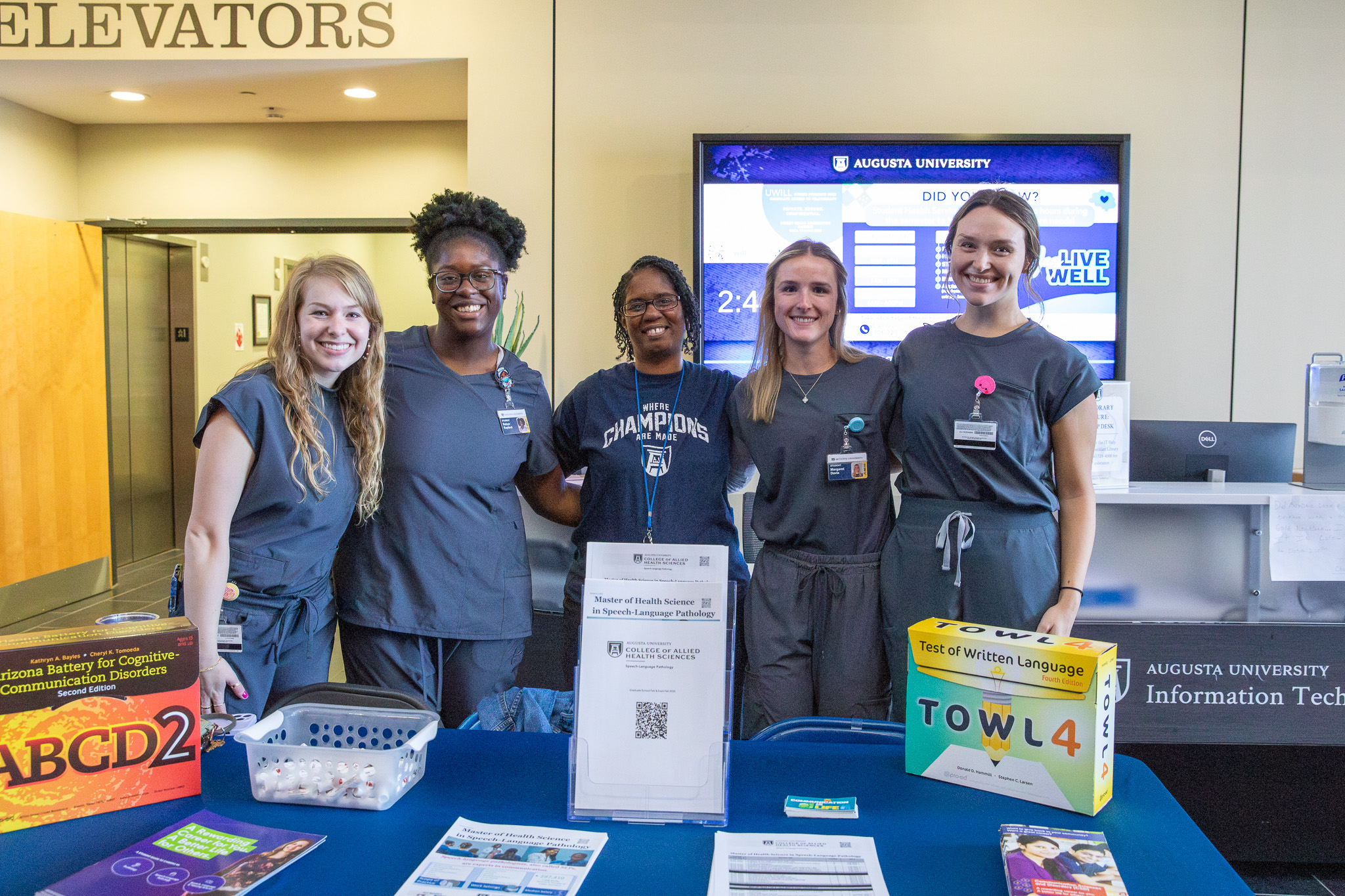 Five individuals wearing blue scrubs smile and stand together behind a table advertising the College of Allied Health Sciences at the 2025 Expo.